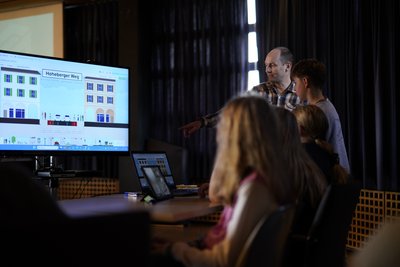 Schüler und Lehrer sehen sich auf IPads und einem Fernseher eine Grafik an