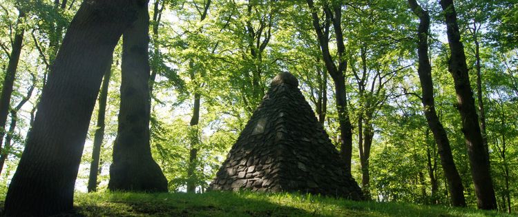 Das Kulturdenkmal Upstalsboom. Eine Pyramide, die aus Steinen aufgebaut wurde. Sie ist von Bäumen umgeben.